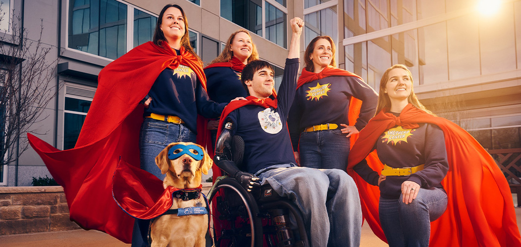 A group of five people and a dog pose confidently in superhero outfits with red capes. One person is in a wheelchair, raising a fist, while a service dog sits alertly at the front. They're outside a modern building with sunlight in the background.