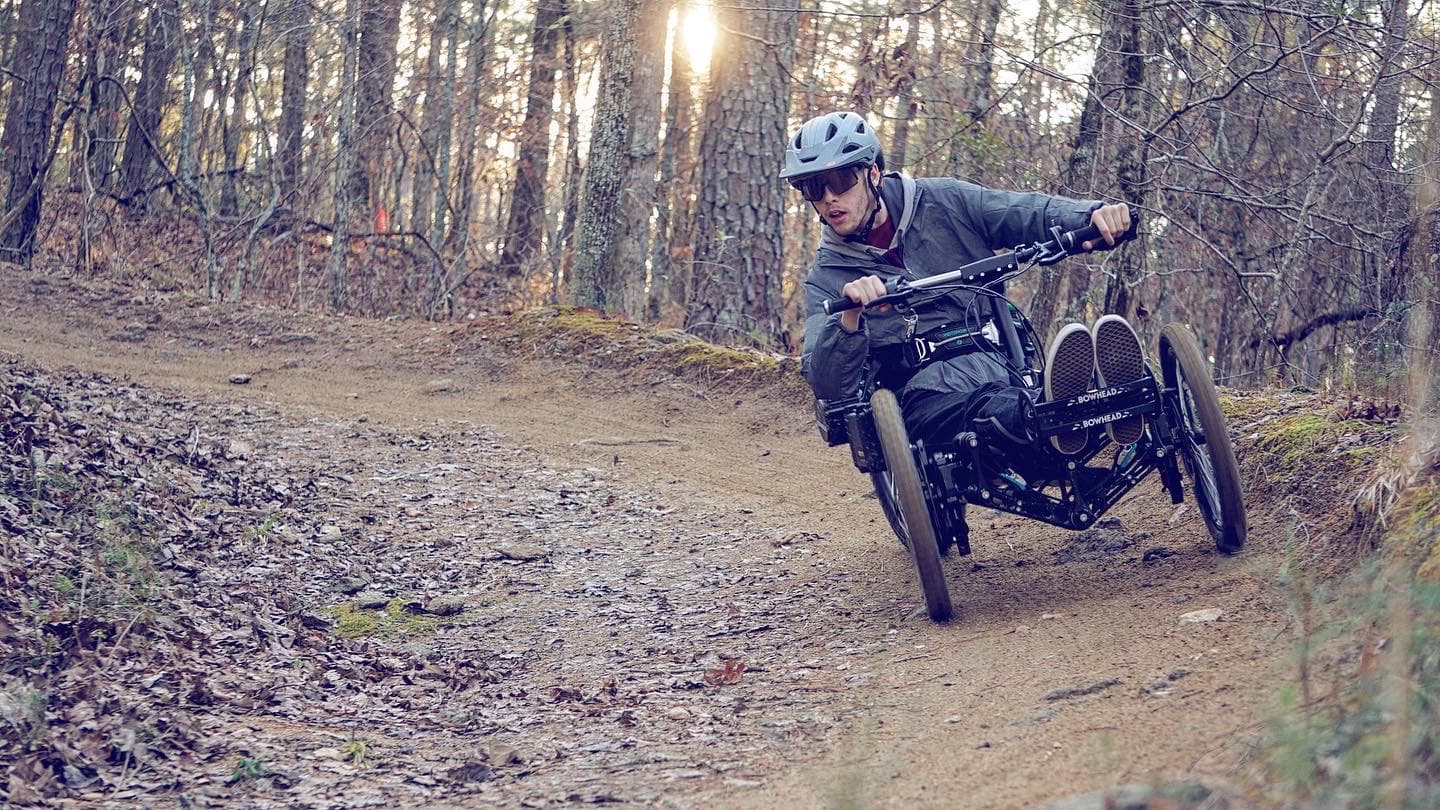 A person wearing a helmet and protective gear rides a four-wheeled off-road adaptive bike on a dirt path through a forest. They lean into the curve, showcasing dynamic motion. Sunlight filters through the trees, highlighting fallen leaves on the path.